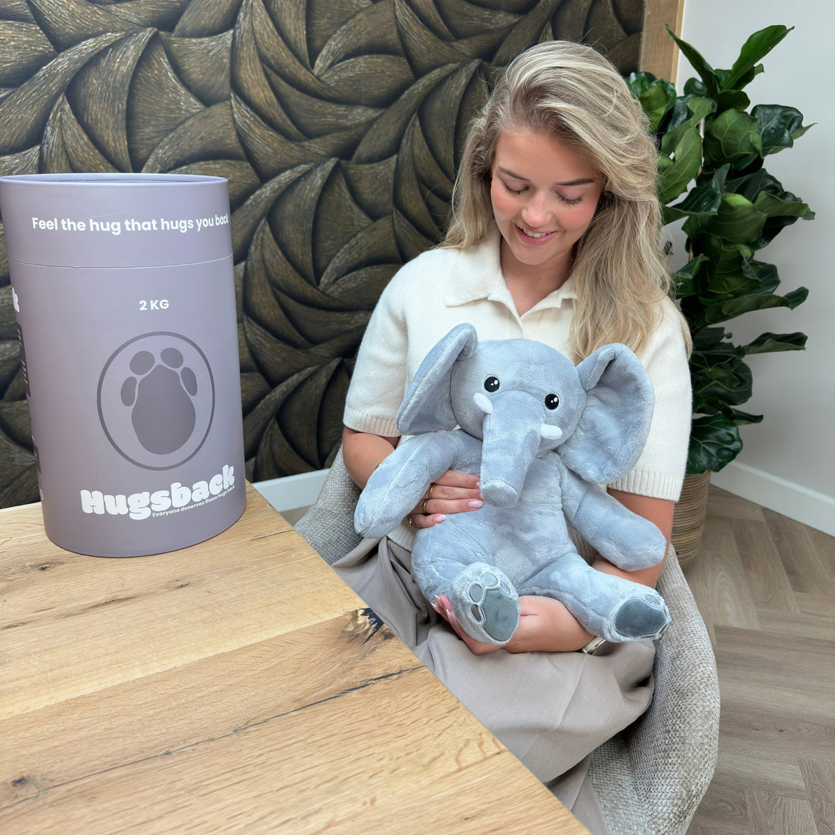 Jonge vrouw in een stoel houdt de Hugsback Olifant op schoot met de grijze cadeaukoker erachter