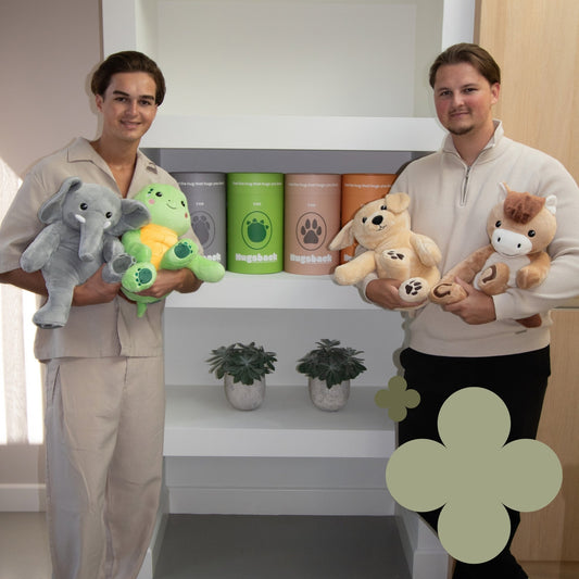 Die Hugsback-Gründer Timo und Noah posieren mit den vier Gewichtskuscheltieren vor einem weißen Regal mit Geschenkröhren und Pflanzen