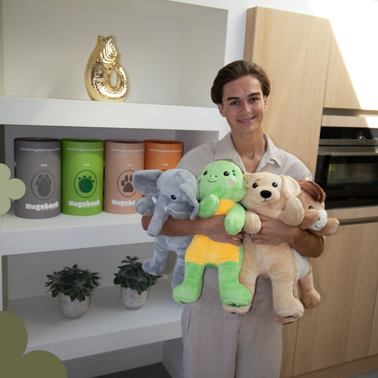 Young man carrying the Hugsback elephant, turtle, dog and horse in his arms in front of a shelf with gift tubes