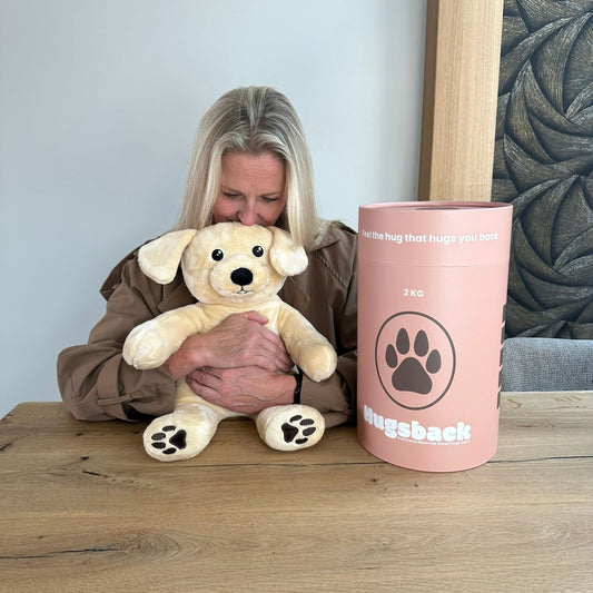 Vrouw knuffelt de beige Hugsback verzwaringsknuffel hond aan tafel naast de zalmroze cadeaukoker