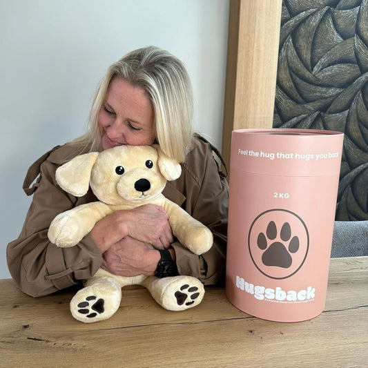 Woman hugging the beige Hugsback weighted plush dog next to the salmon-pink gift tube
