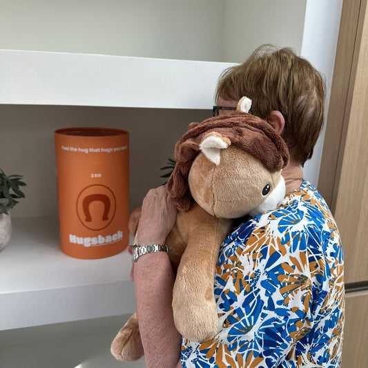 Oudere vrouw knuffelt de bruine Hugsback verzwaringsknuffel paard naast de oranje cadeaukoker