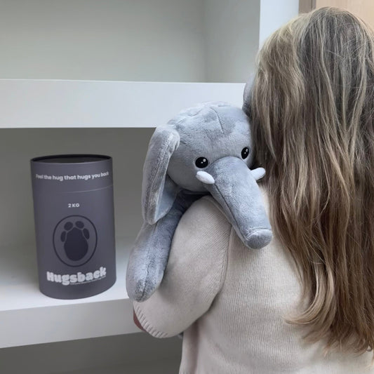Giovane donna davanti a un armadio bianco che porta l'elefante Hugsback sulla spalla con il tubo regalo grigio alle spalle