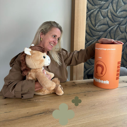 Femme souriante avec la peluche lestée cheval Hugsback marron sur les genoux à table en attrapant le tube cadeau orange