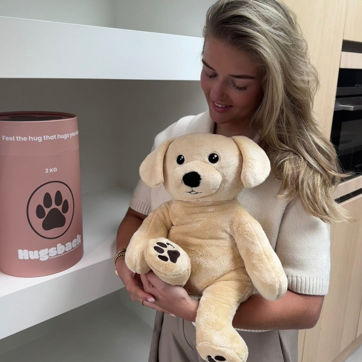 Vrouw houdt de beige Hugsback verzwaringsknuffel hond vast naast de zalmroze cadeaukoker