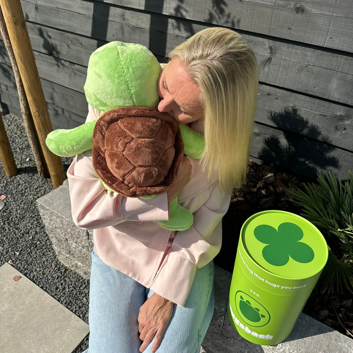 Vrouw knuffelt buiten de groene Hugsback Schildpad naast de groene cadeaukoker