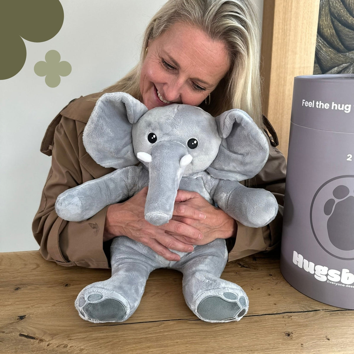 Femme souriante serrant la peluche lestée éléphant Hugsback grise dans ses bras à table à côté du tube cadeau gris