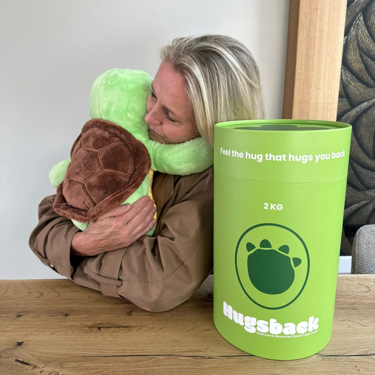 Vrouw knuffelt de groene Hugsback verzwaringsknuffel schildpad aan tafel naast de groene cadeaukoker