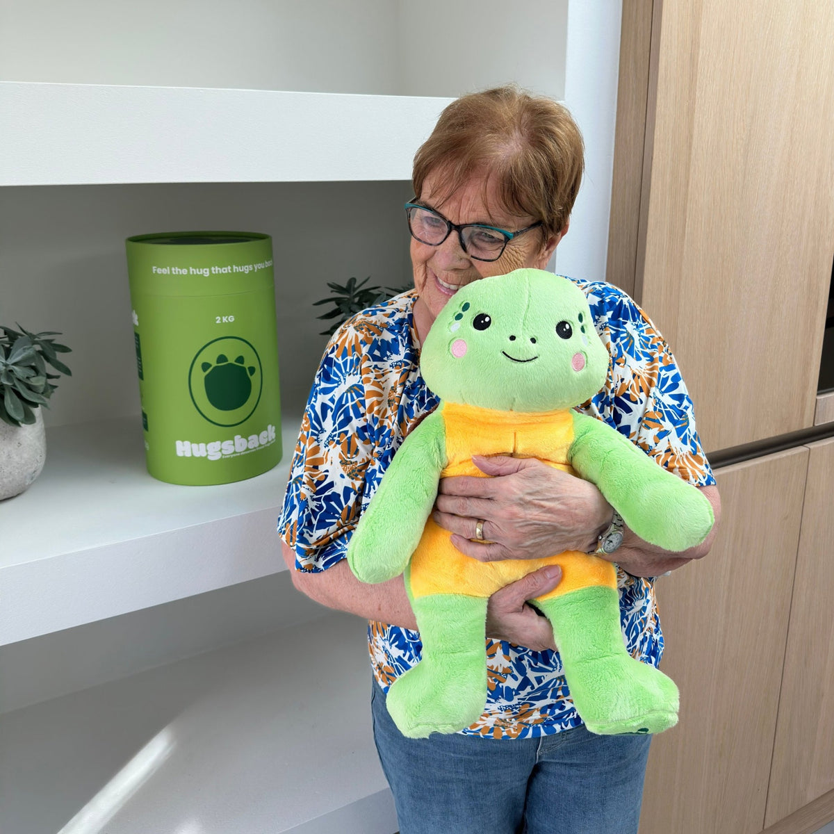 Older woman holding the green Hugsback weighted plush turtle next to the green gift tube