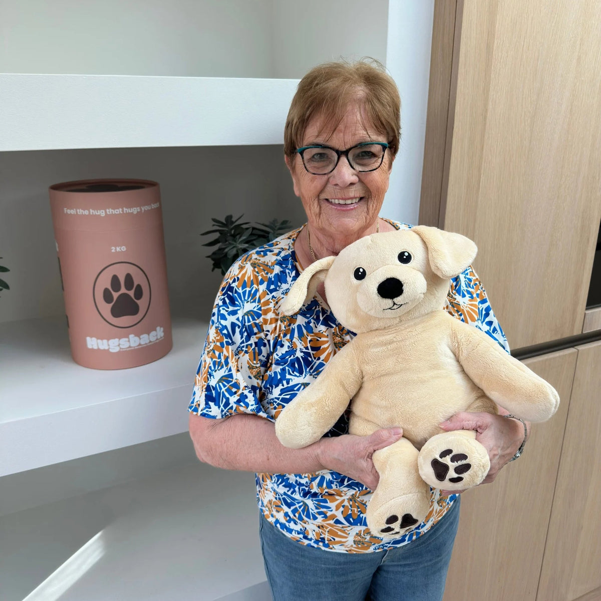 Older woman with the beige Hugsback weighted plush dog next to the salmon-pink gift tube