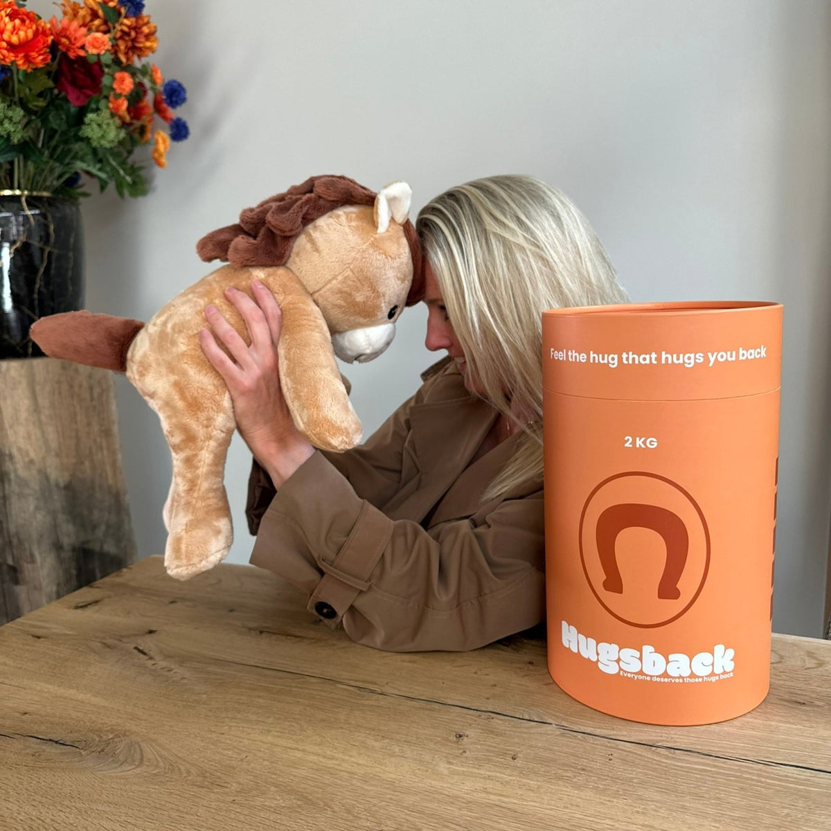 Vrouw houdt de bruine Hugsback verzwaringsknuffel paard omhoog aan tafel naast de oranje cadeaukoker