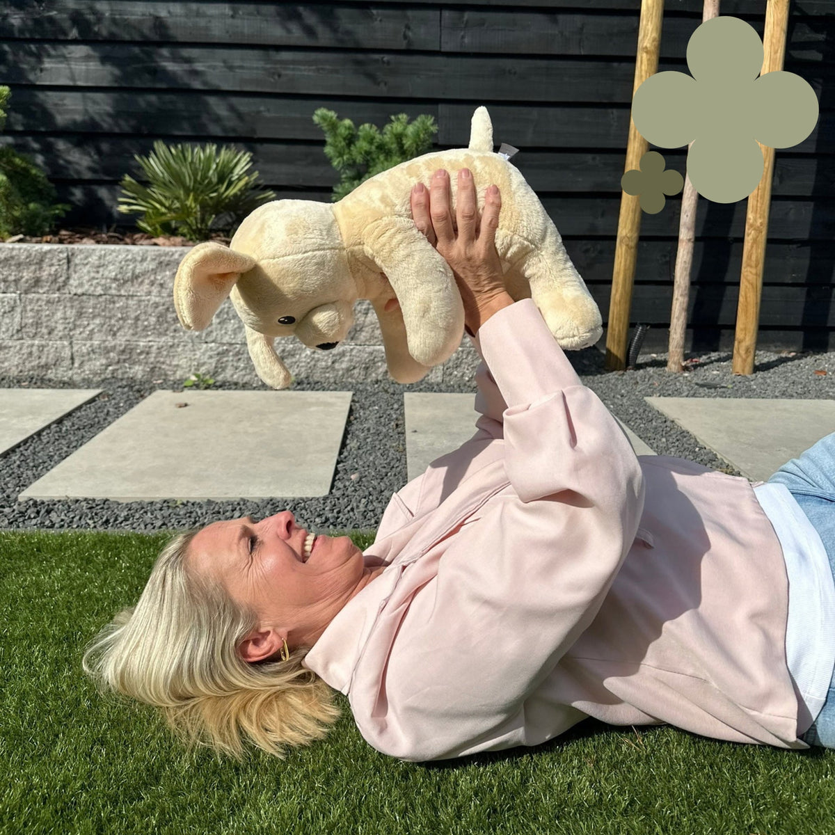 Femme allongée dans l'herbe soulevant la peluche lestée chien Hugsback beige dans le jardin