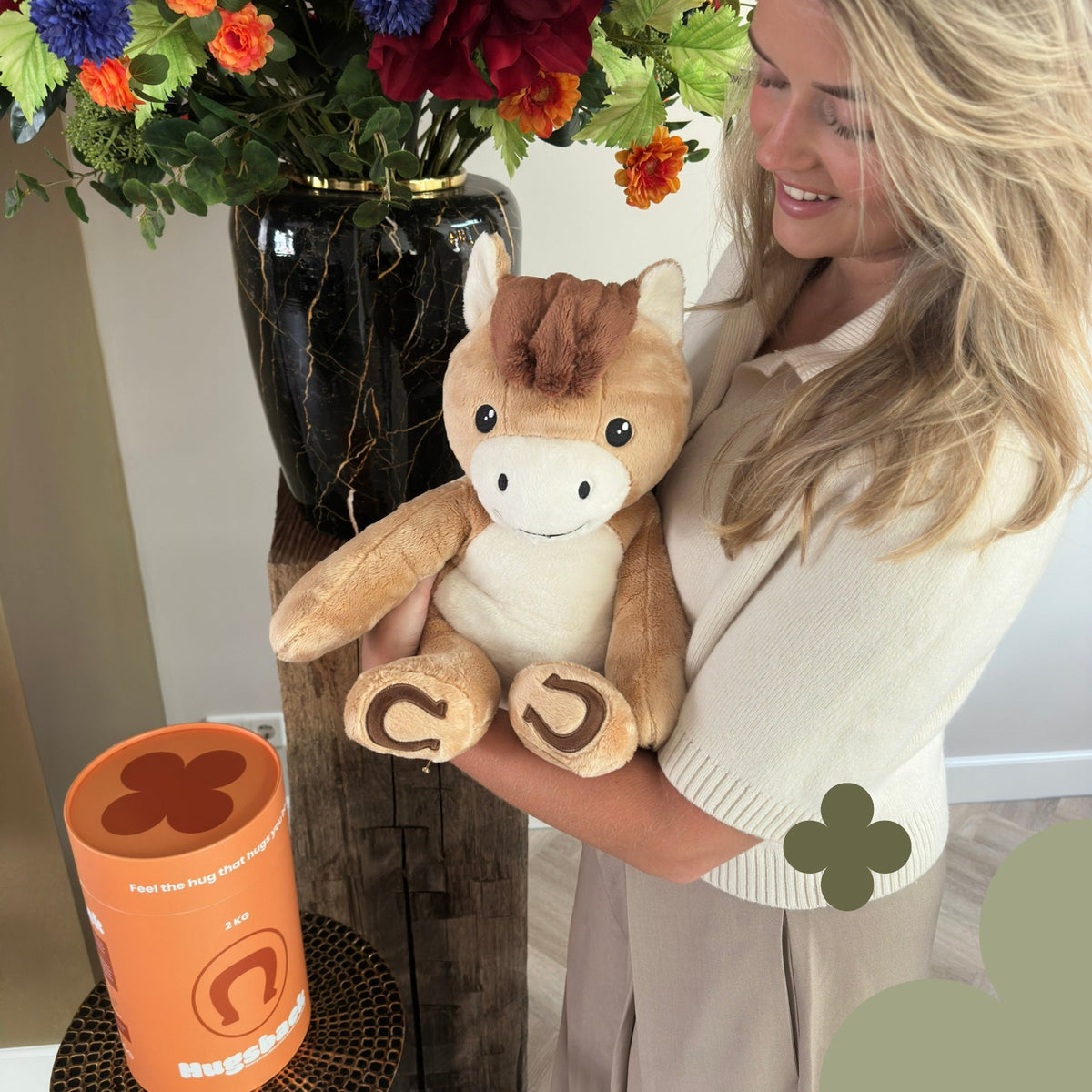 Woman holding the brown Hugsback weighted plush horse beside a bouquet of flowers next to the orange gift tube