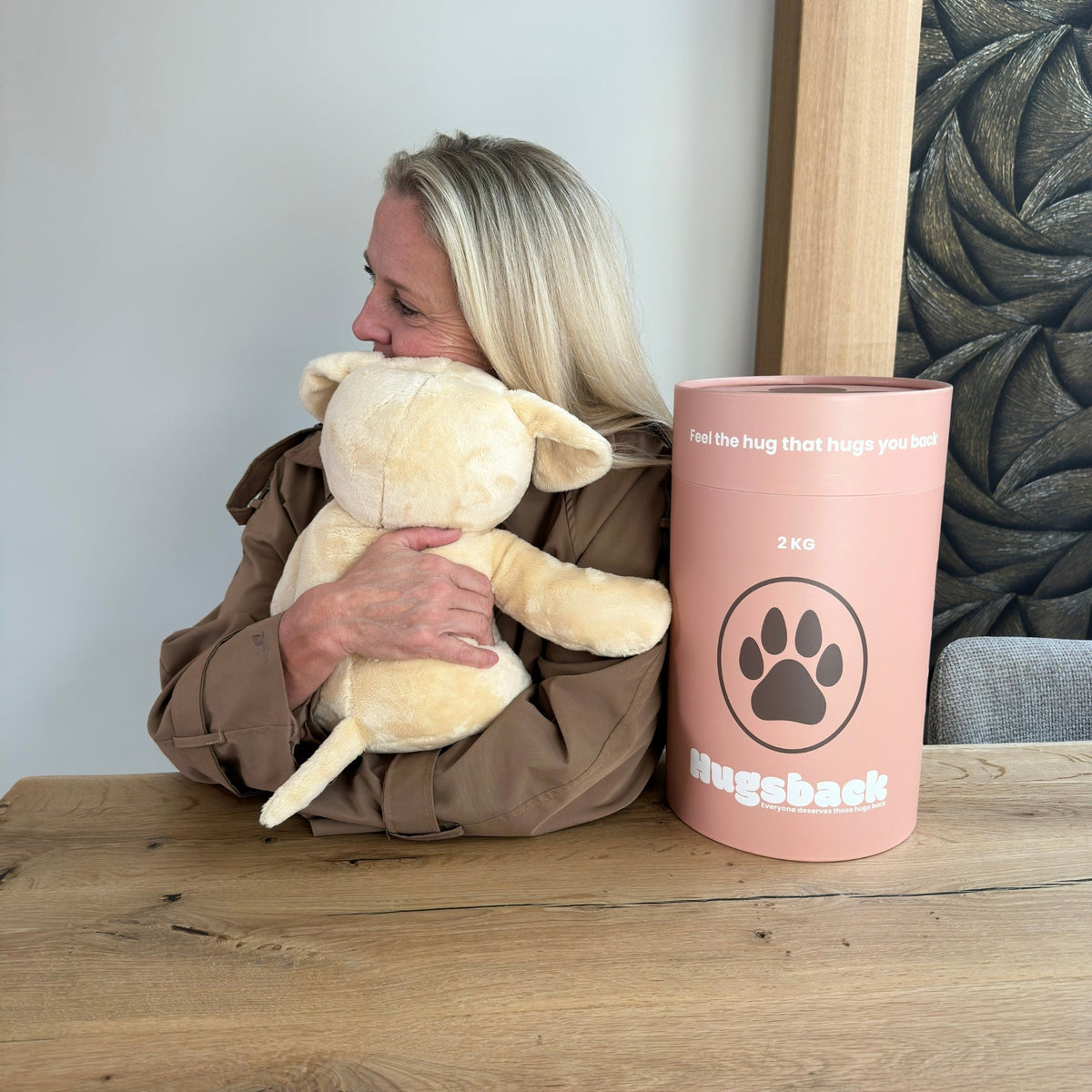Woman hugging the beige Hugsback weighted plush dog at a table with her face pressed against it