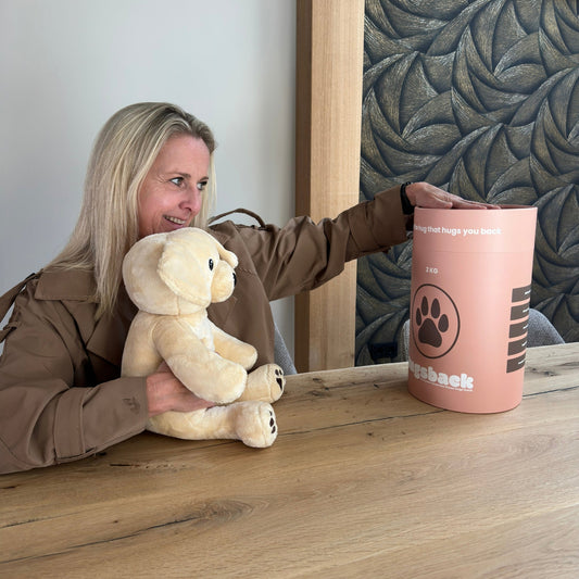 Vrouw houdt de beige Hugsback verzwaringsknuffel hond vast aan tafel en pakt de zalmroze cadeaukoker