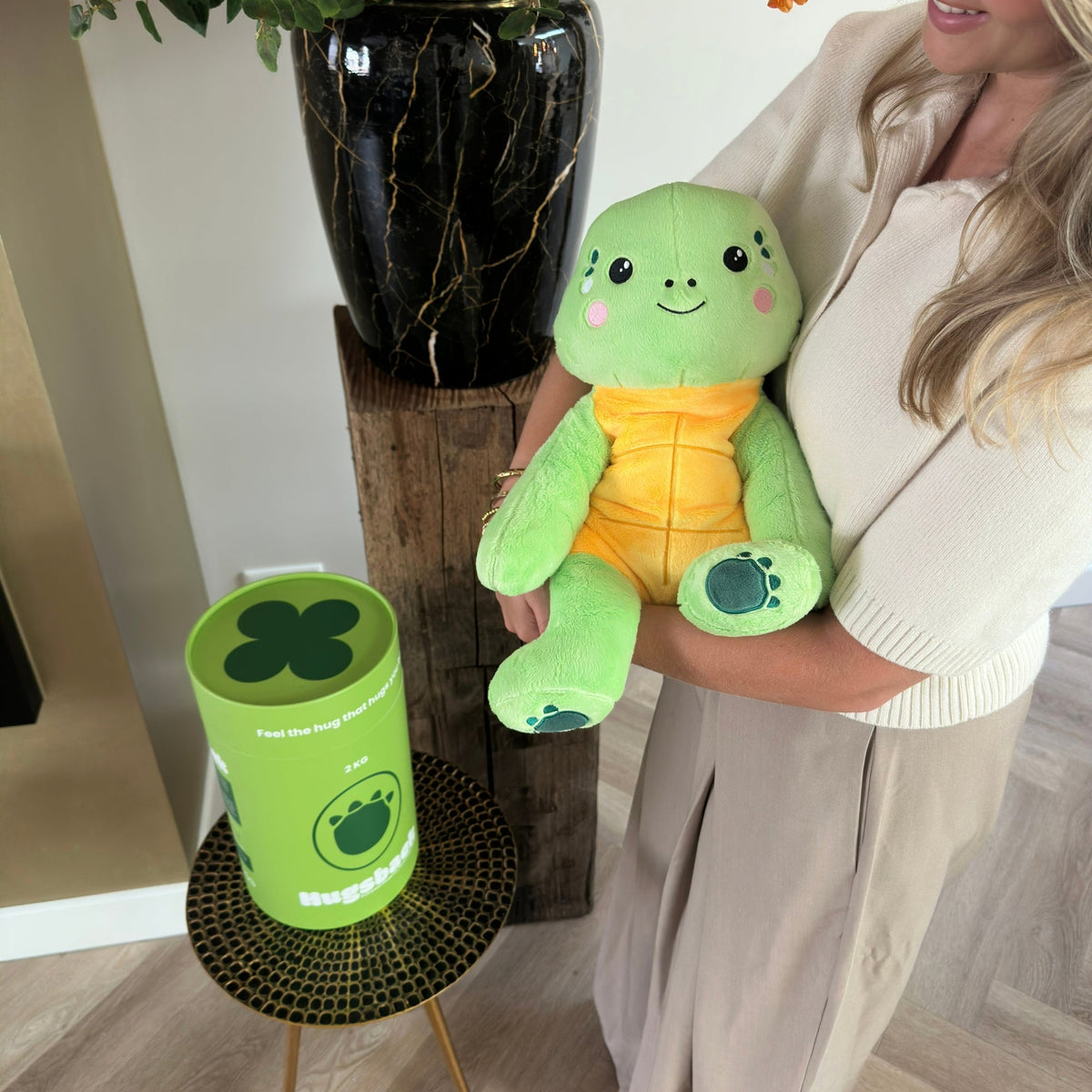 Vrouw houdt de groene Hugsback verzwaringsknuffel schildpad vast bij een boeket bloemen naast de groene cadeaukoker