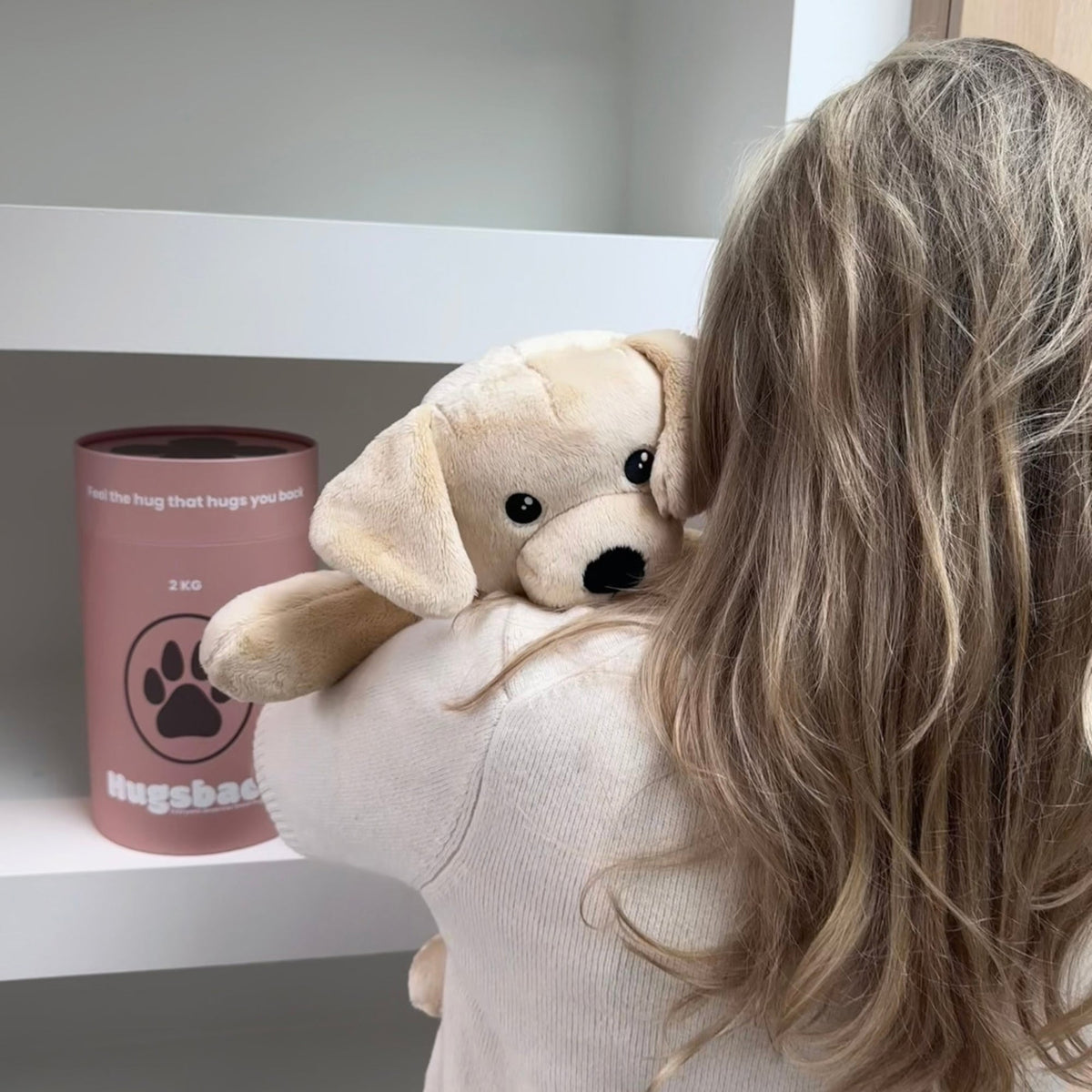 Woman hugging the beige Hugsback weighted plush dog over her shoulder seen from behind next to the salmon-pink gift tube