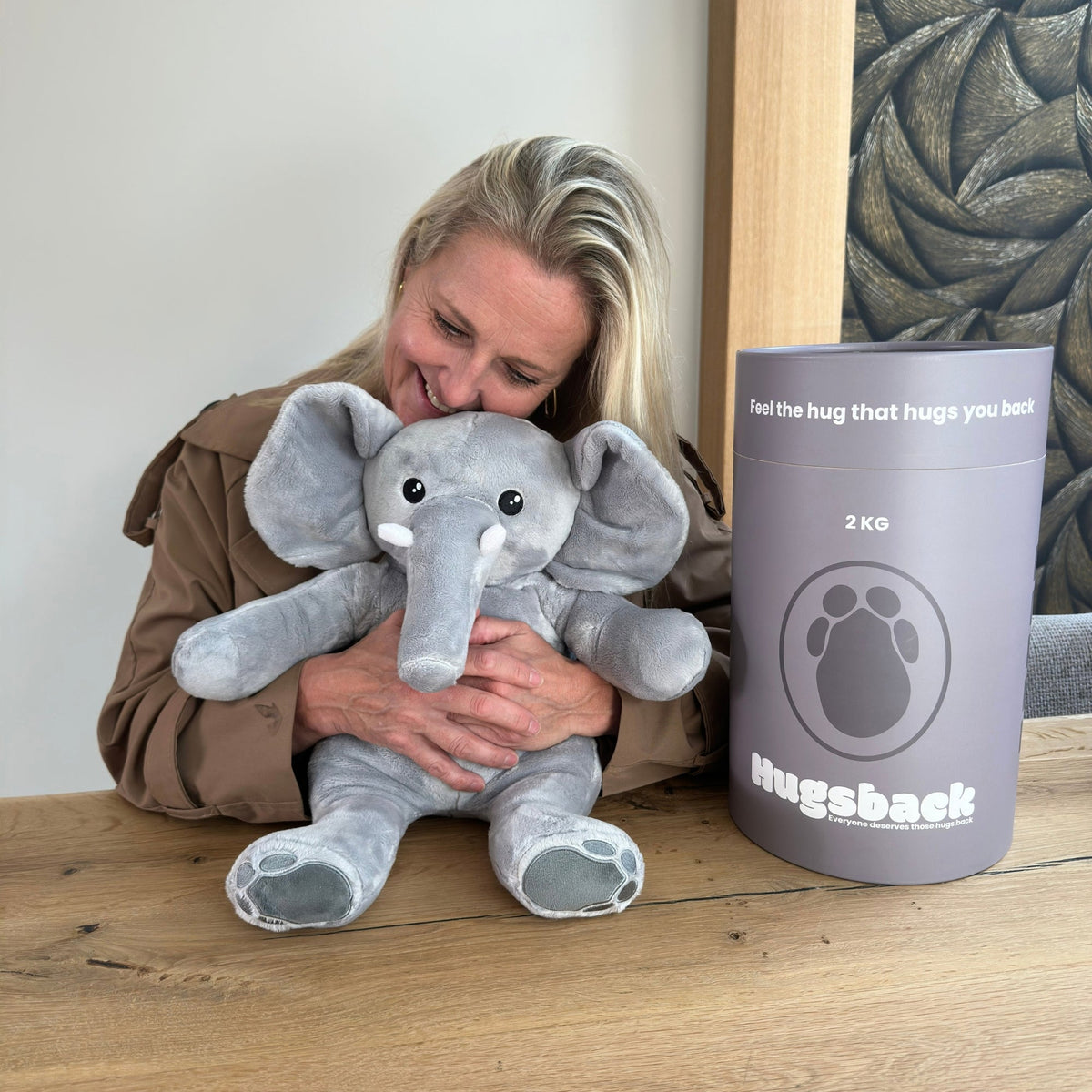 Frau am Tisch kuschelt den Hugsback Elefanten neben der grauen Geschenkröhre