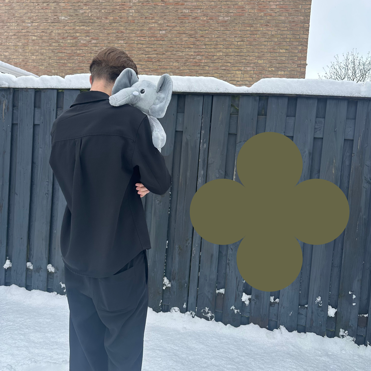 Man met de grijze Hugsback verzwaringsknuffel olifant op zijn schouder bij een schutting in de sneeuw