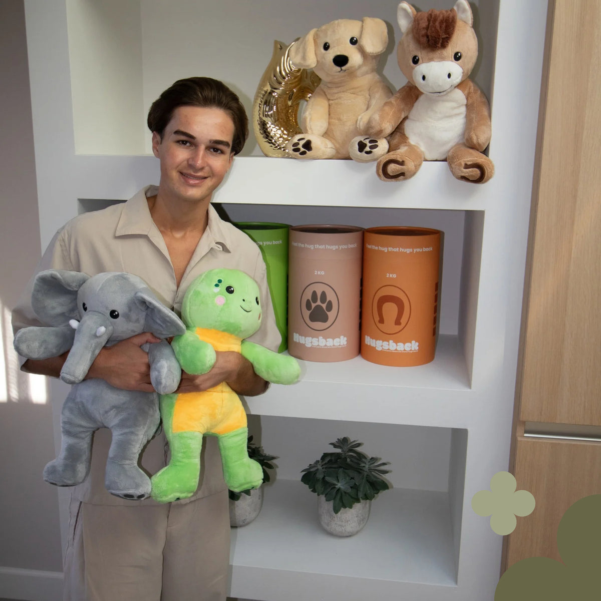 Jeune homme avec les peluches lestées éléphant et tortue devant un placard rempli de tubes cadeaux Hugsback