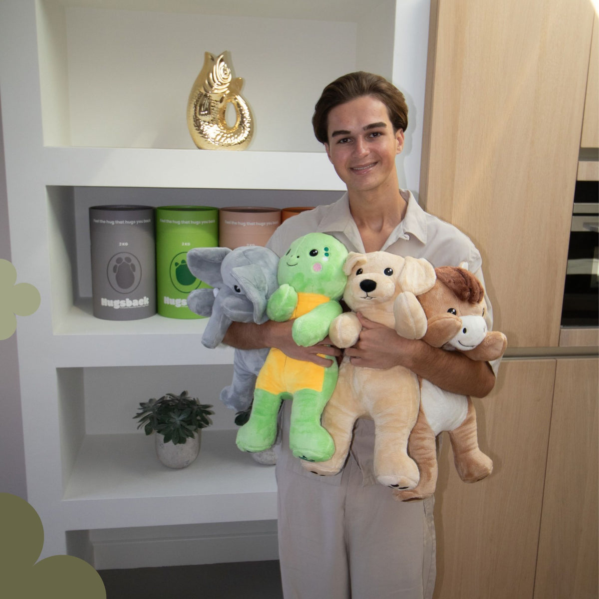 Junger Mann mit allen vier Hugsback Gewichtskuscheltieren in den Armen vor einem Schrank mit Hugsback-Geschenkröhren