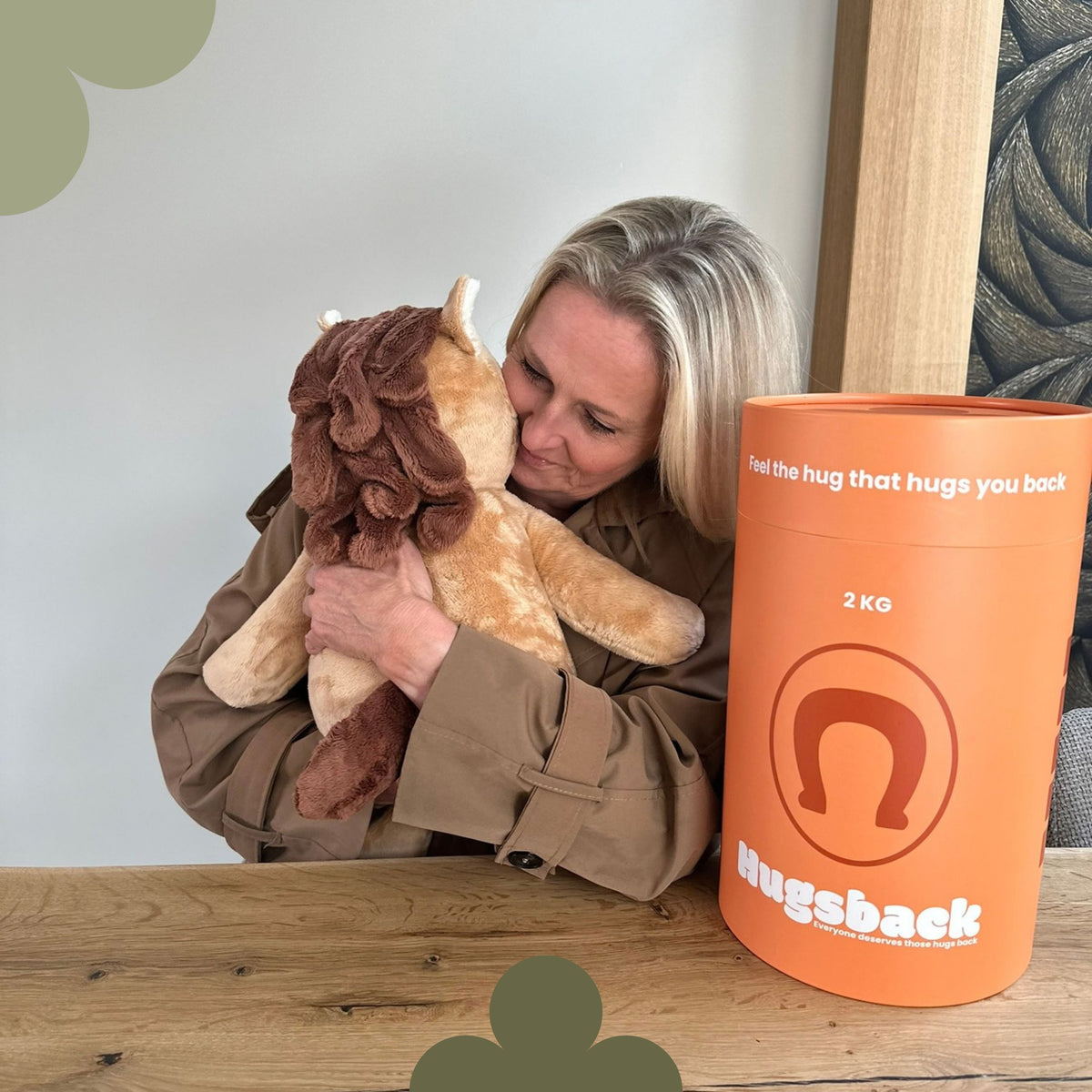 Frau am Tisch, die das braune Gewichtskuscheltier Pferd neben der orangen Hugsback-Geschenkröhre knuddelt