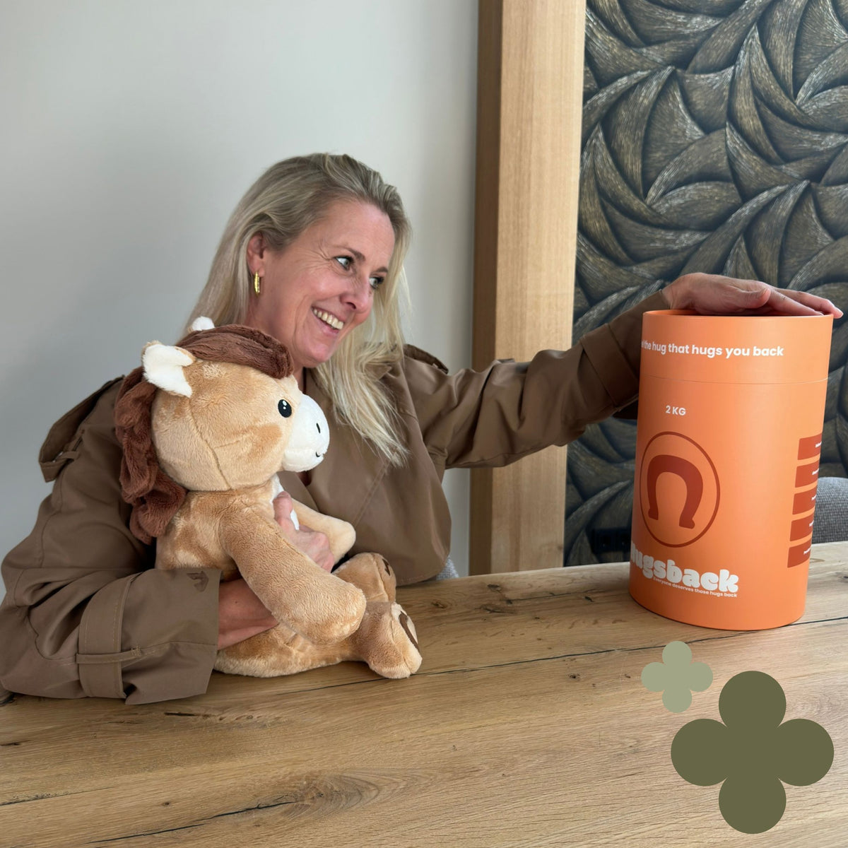 Femme à table qui câline la peluche lestée cheval marron et tient le tube cadeau Hugsback orange