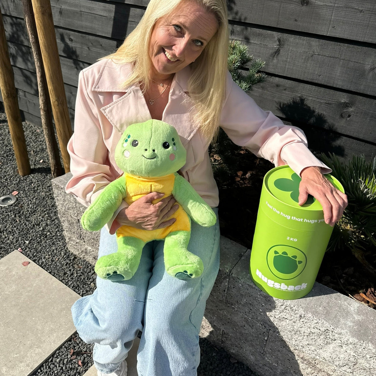 Femme assise dehors avec la peluche lestée tortue verte à côté du tube cadeau Hugsback vert