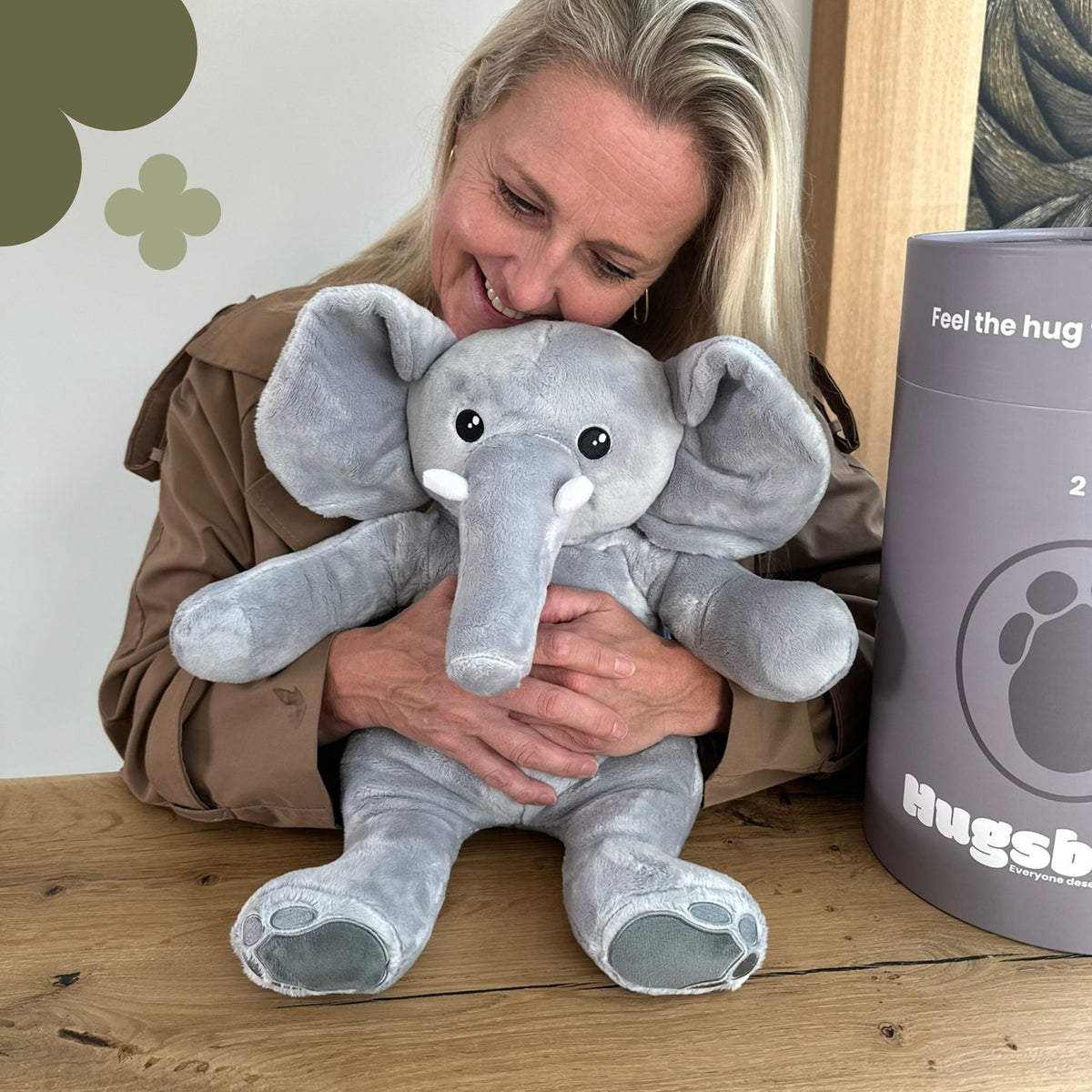 Femme riant à table qui câline la peluche lestée éléphant grise à côté du tube cadeau gris