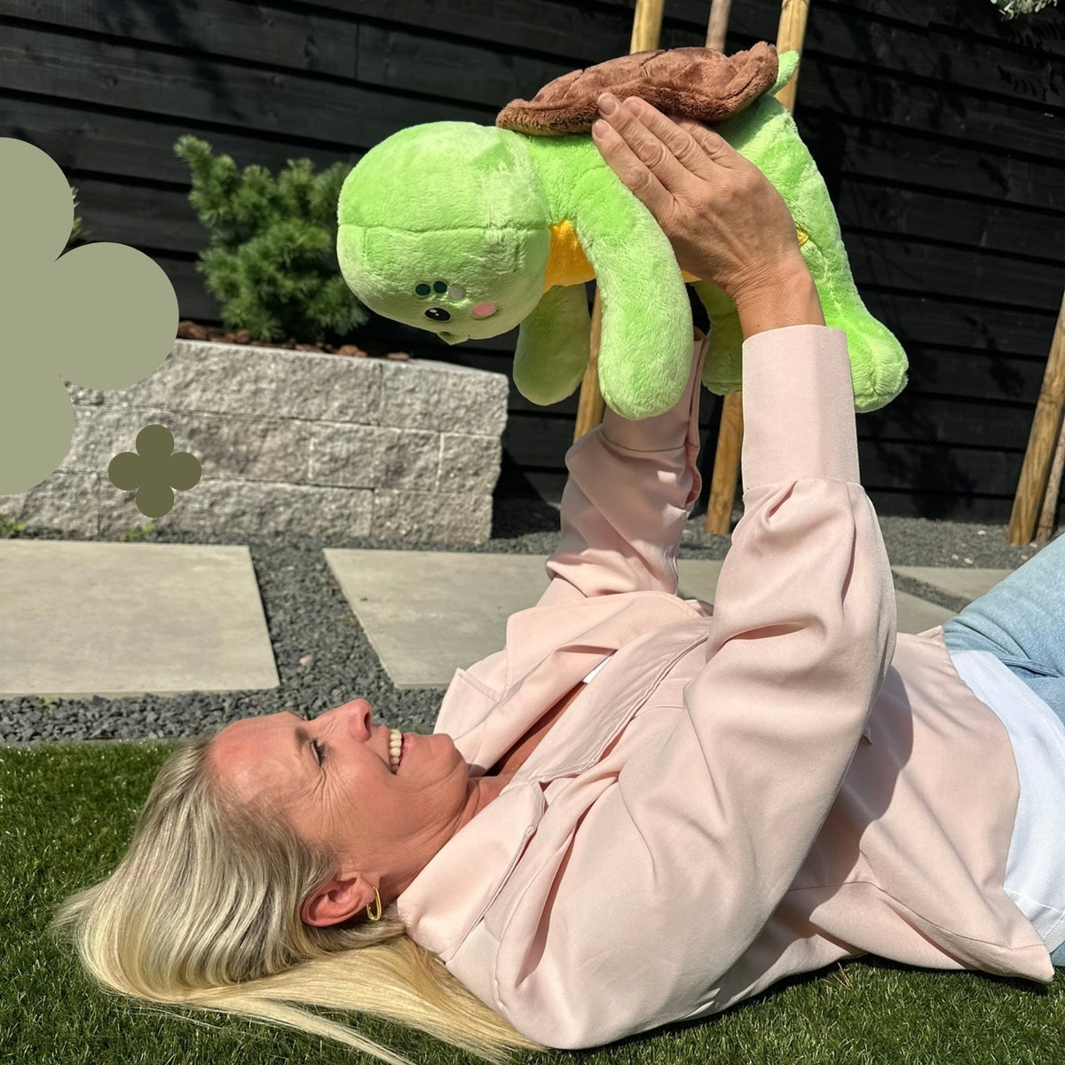 Frau, die im Gras liegt und das grüne Gewichtskuscheltier Schildkröte hochhält