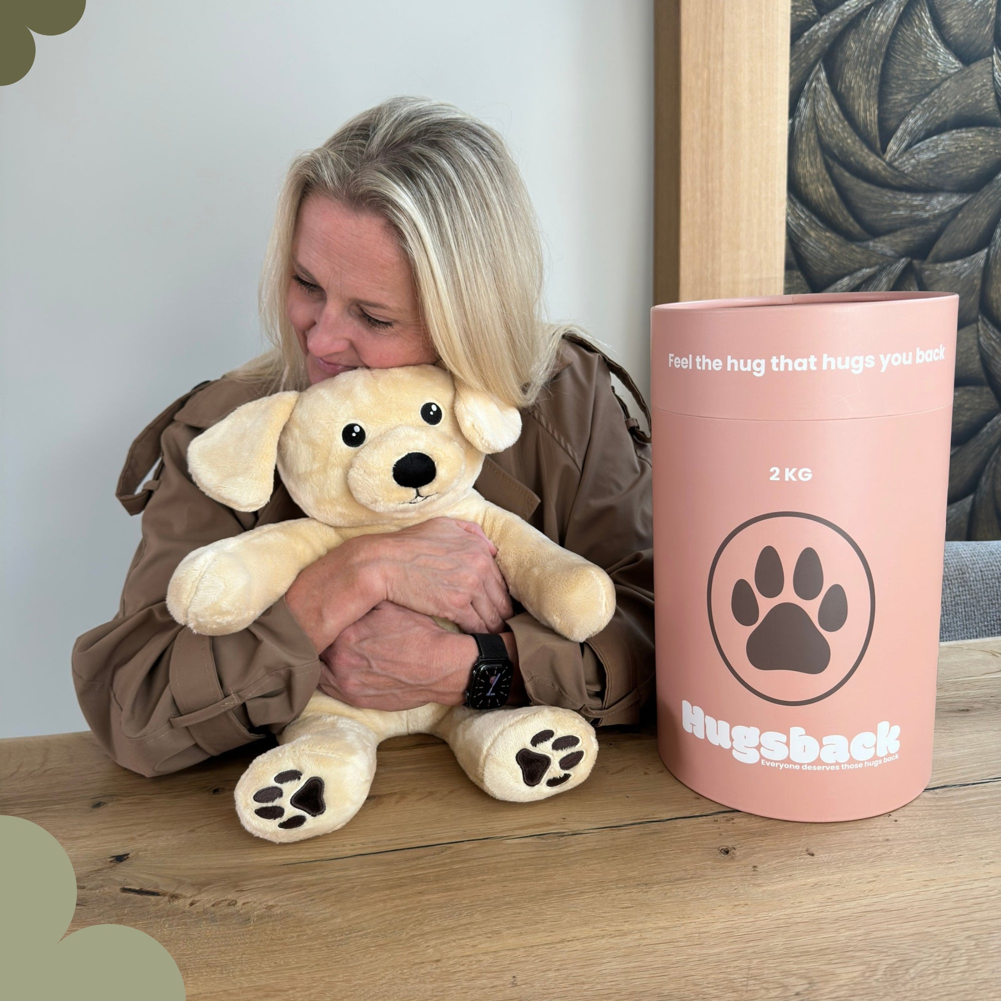 Vrouw aan tafel die de beige verzwaringsknuffel hond knuffelt naast de roze Hugsback cadeaukoker