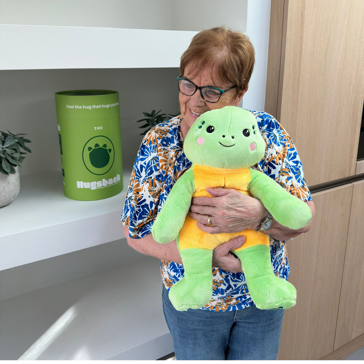 Ältere Frau mit dem grünen Gewichtskuscheltier Schildkröte vor einem Schrank mit Hugsback-Geschenkröhren