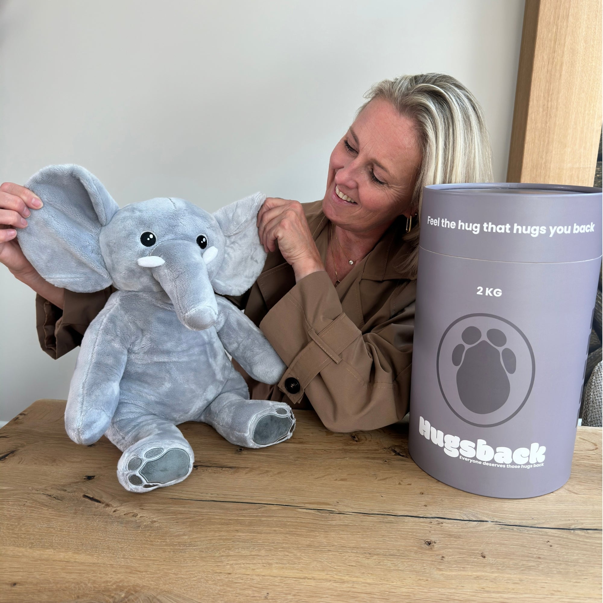 Vrouw aan tafel met de grijze verzwaringsknuffel olifant naast de grijze Hugsback cadeaukoker