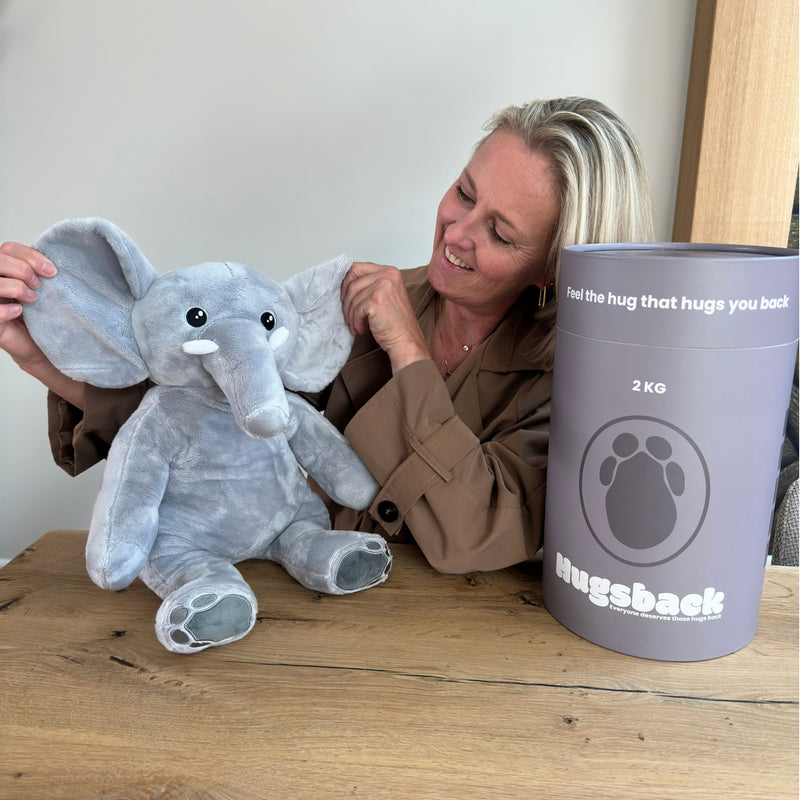 Vrouw aan tafel met de grijze verzwaringsknuffel olifant naast de grijze Hugsback cadeaukoker