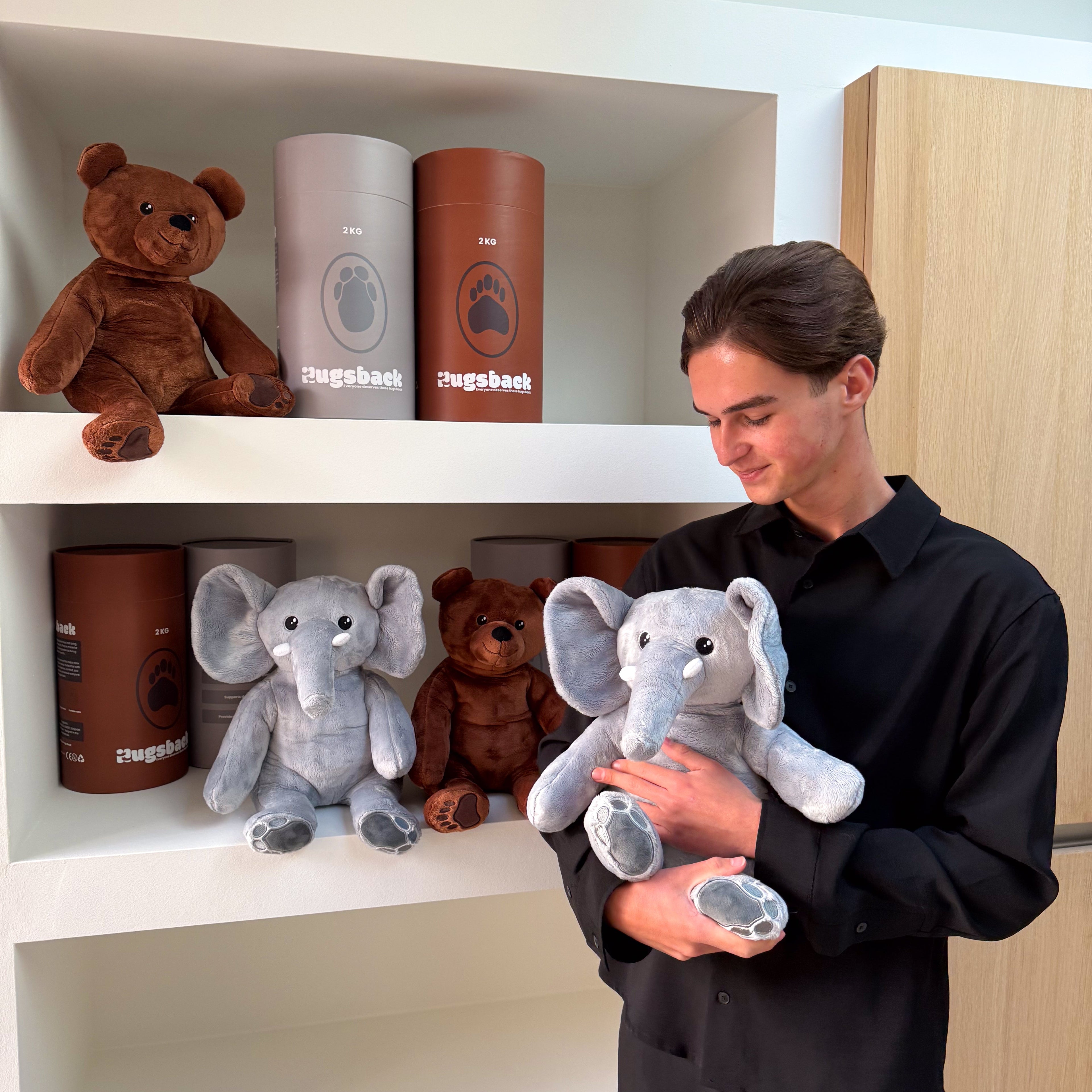Man holding a plush elephant toy in front of shelves with teddy bears and cylindrical containers.