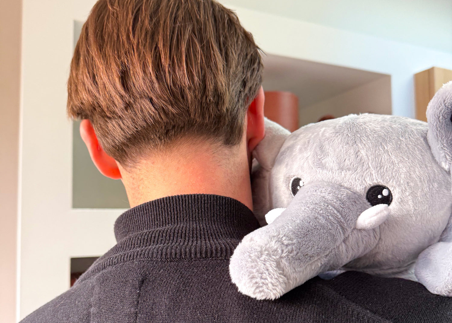 Timo knuffelt de Hugsback verzwaringsolifant over zijn schouder, met een rustige en warme uitstraling voor de wandkast vol Hugsback knuffels en kokers.