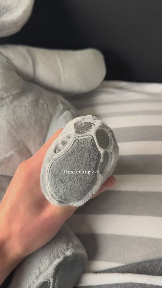 Duitstalige video van een hand die het pootje van de grijze verzwaringsknuffel olifant vasthoudt op een gestreept bed